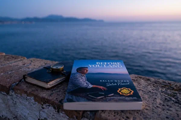 A physical copy of Before You Land resting on a weathered stone terrace at blue hour, beside a black leather notebook and an antique brass compass, with the Mediterranean Sea stretching to the horizon.