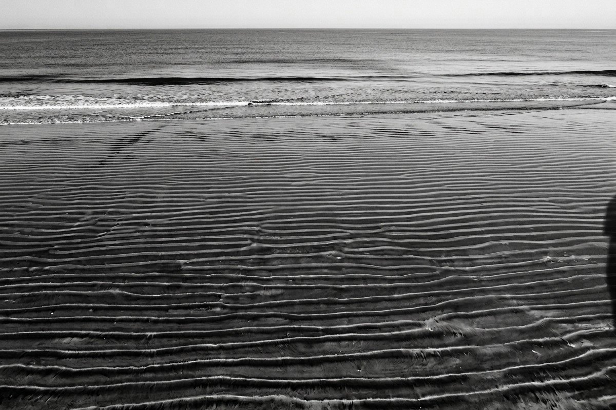 Aerial black and white photograph of Pedregalejo beach at low tide, parallel lines in sand formed by waves, a subtle human shadow at the frame’s edge – evoking purposeful migration.