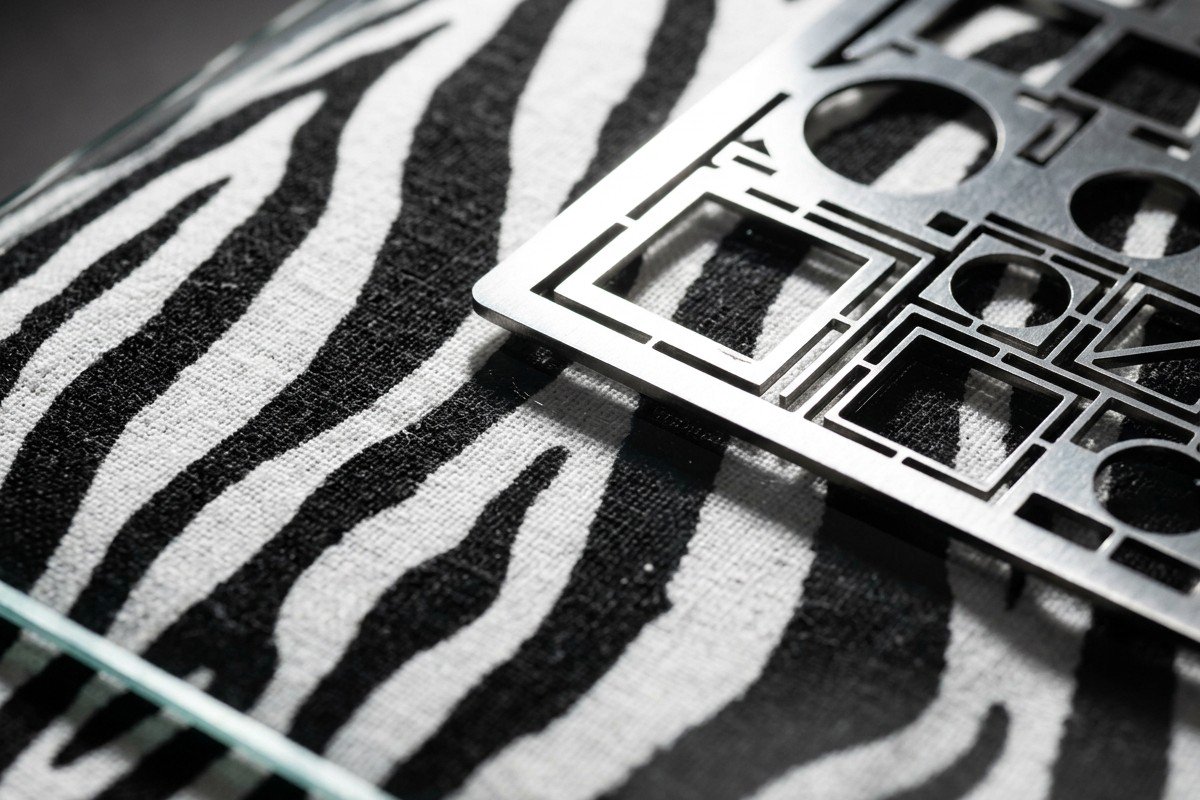 Extreme close-up of black and white zebra-striped fabric on glass with a metal stencil resting on it, emphasizing texture and contrast – identity as a signal.