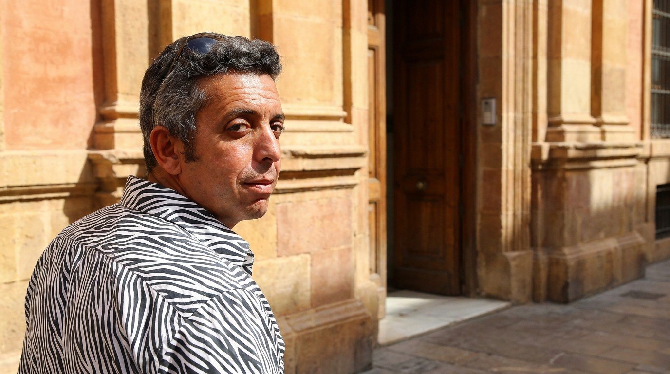 Salah Nomad in his zebra-striped shirt standing before the closed wooden door of an old Andalusian bank in harsh midday light, his face turned in three-quarter profile toward the door, his shadow stretching on the pavement.