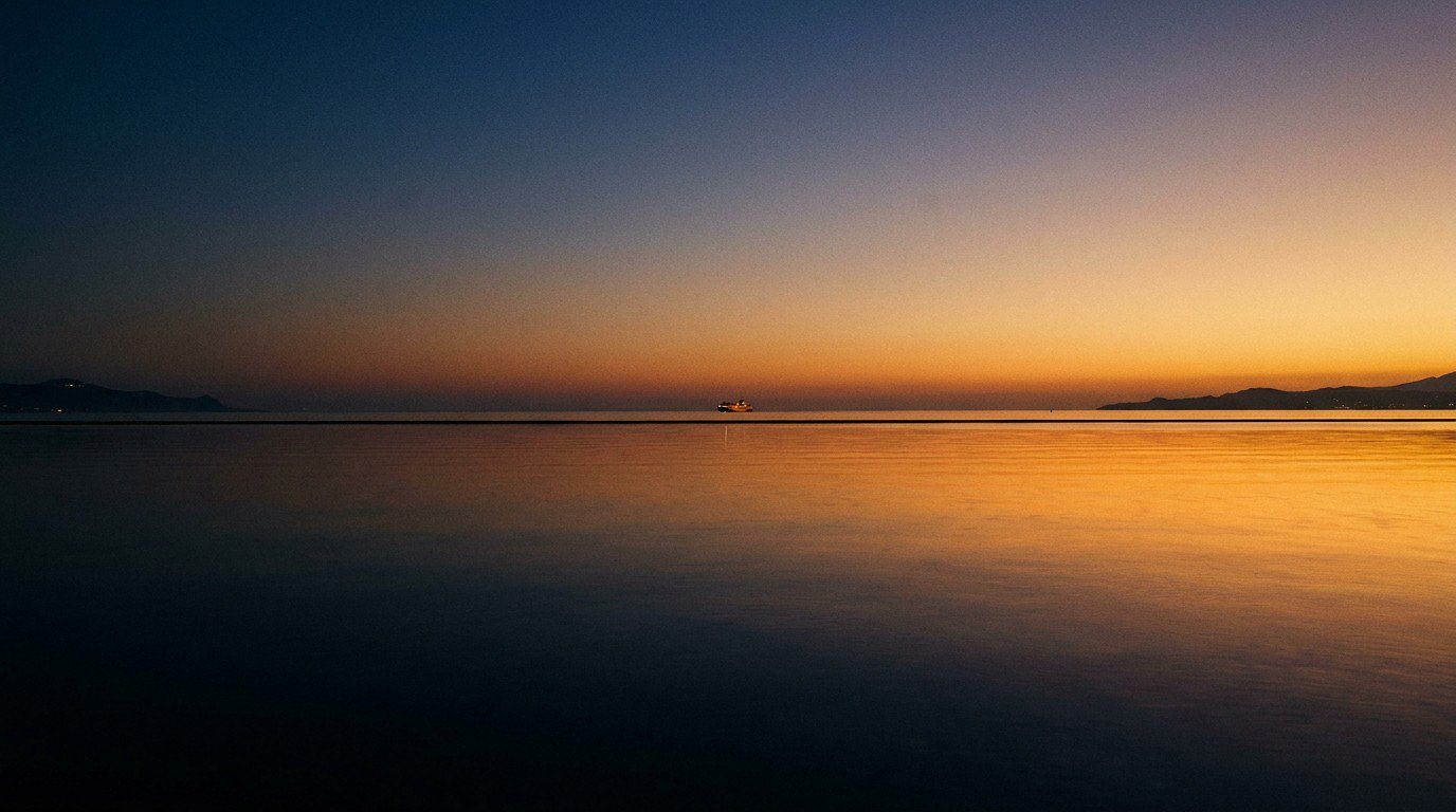The Strait of Gibraltar at dusk, a single ferry far in the distance, a small point of light on the water, the two coastlines of Tarifa and Tangier faintly visible on either side, the sea almost oil-flat reflecting the last amber light beneath a sky graduating from amber to deep indigo.