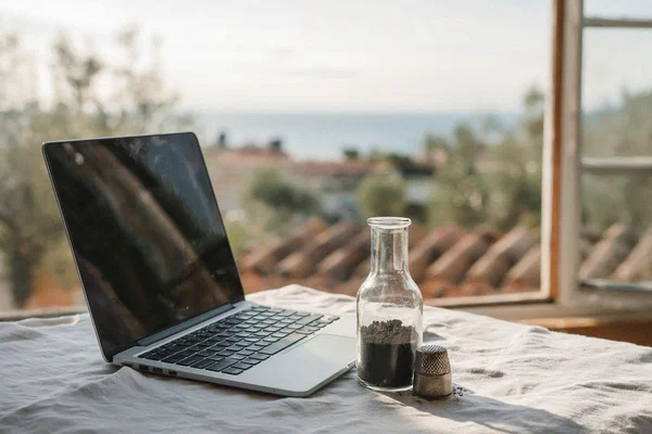 A minimalist nomadic workspace with a laptop, a small glass vial of soil, and an ancient silver thimble on white linen, overlooking a sun-drenched Mediterranean coastline with olive trees.